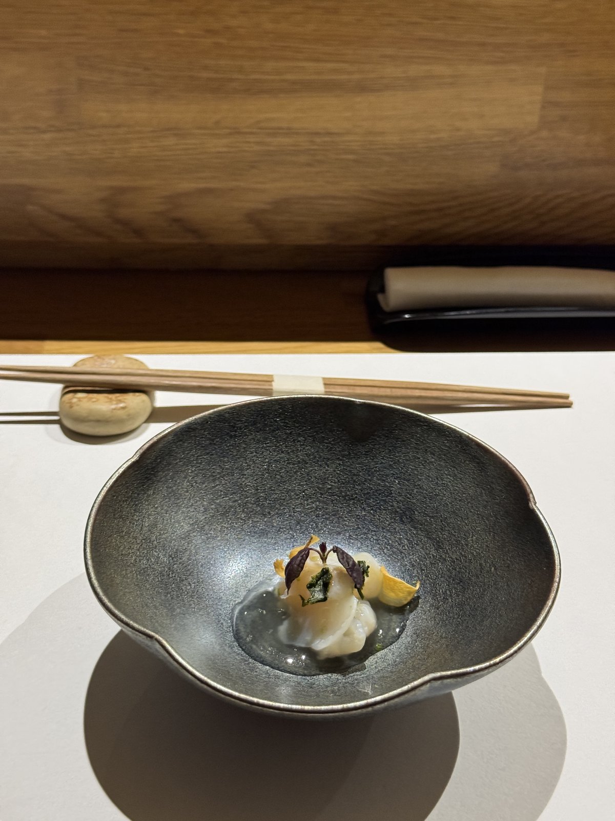 A single scallop dressed with shiso leaves and edible flowers, in a dark ceramic bowl on a counter, chopsticks resting above.